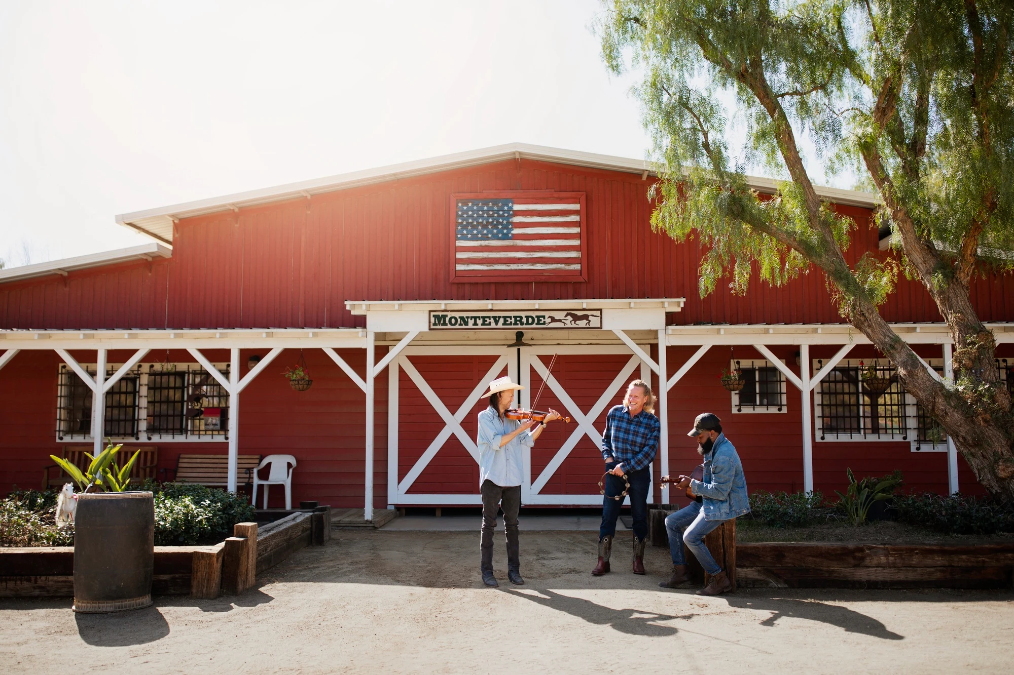 Red barn-style barndominium with white trim and an American flag, featuring people playing musical instruments in a sunny outdoor setting at Monteverde.