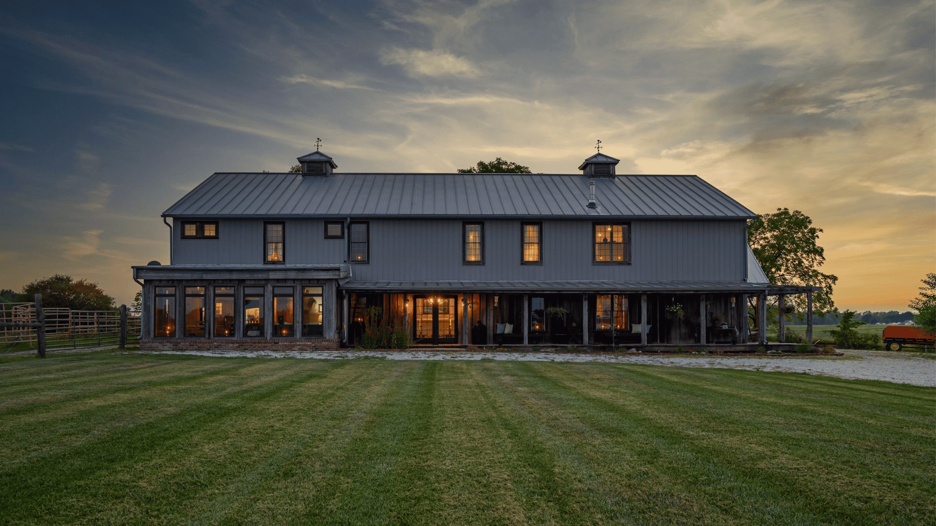 Large two-story barndominium with gray metal siding and roof, wraparound porch, and warm glowing windows at sunset, set on a spacious green lawn.