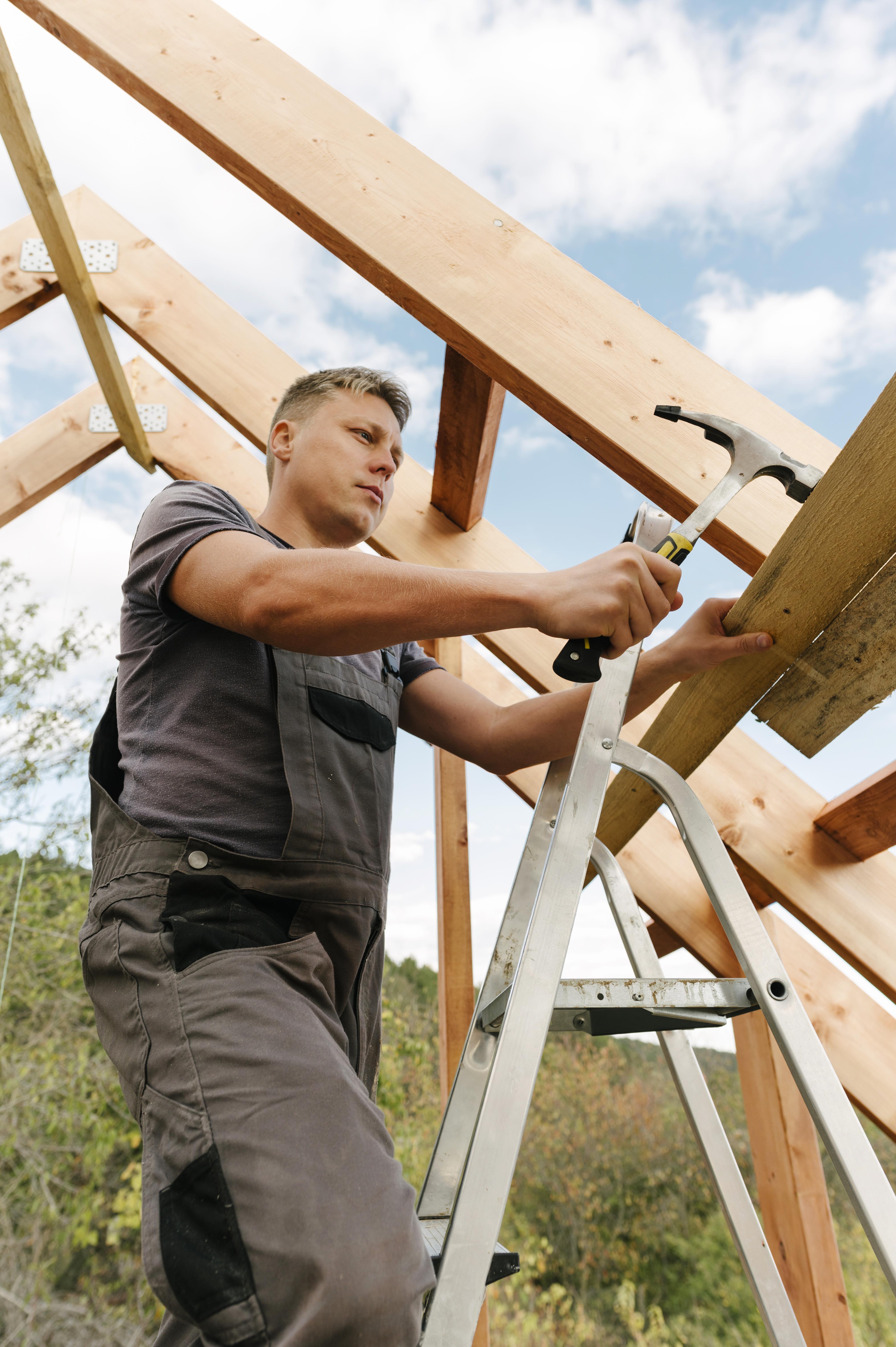 Construction worker on a ladder installing wooden beams for a new barndominium roof frame, working outdoors under a blue sky.